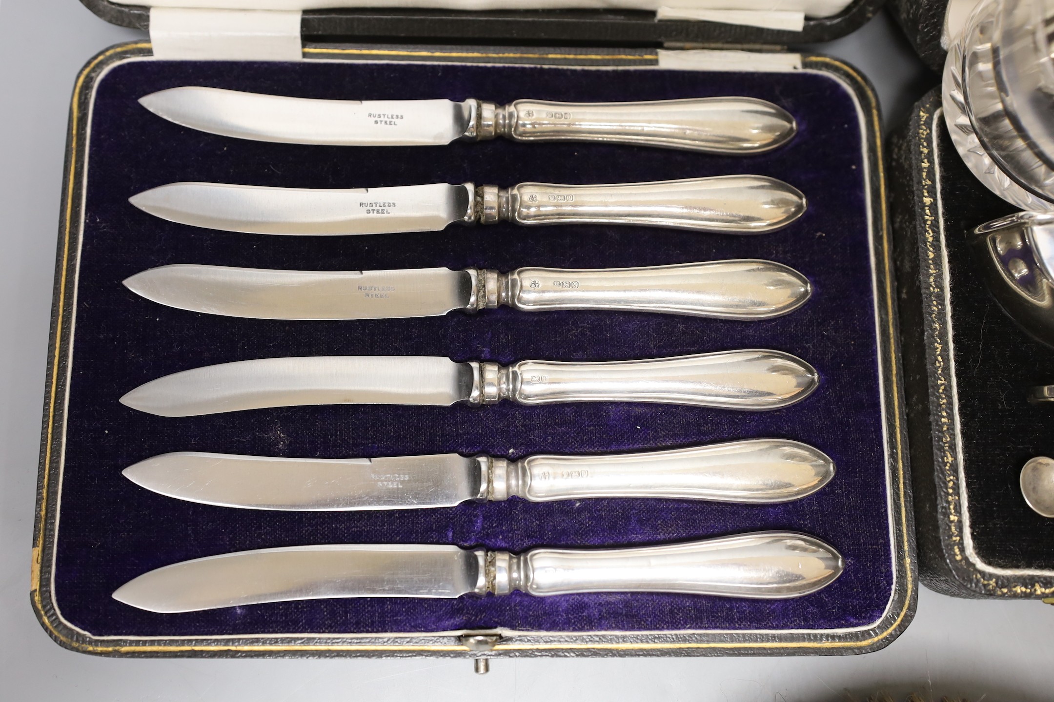 A cased 1950's silver three piece condiment set, two cased sets of six silver handled tea knives, a silver mounted glass salts bottle, silver mounted hand mirror and two brushes.